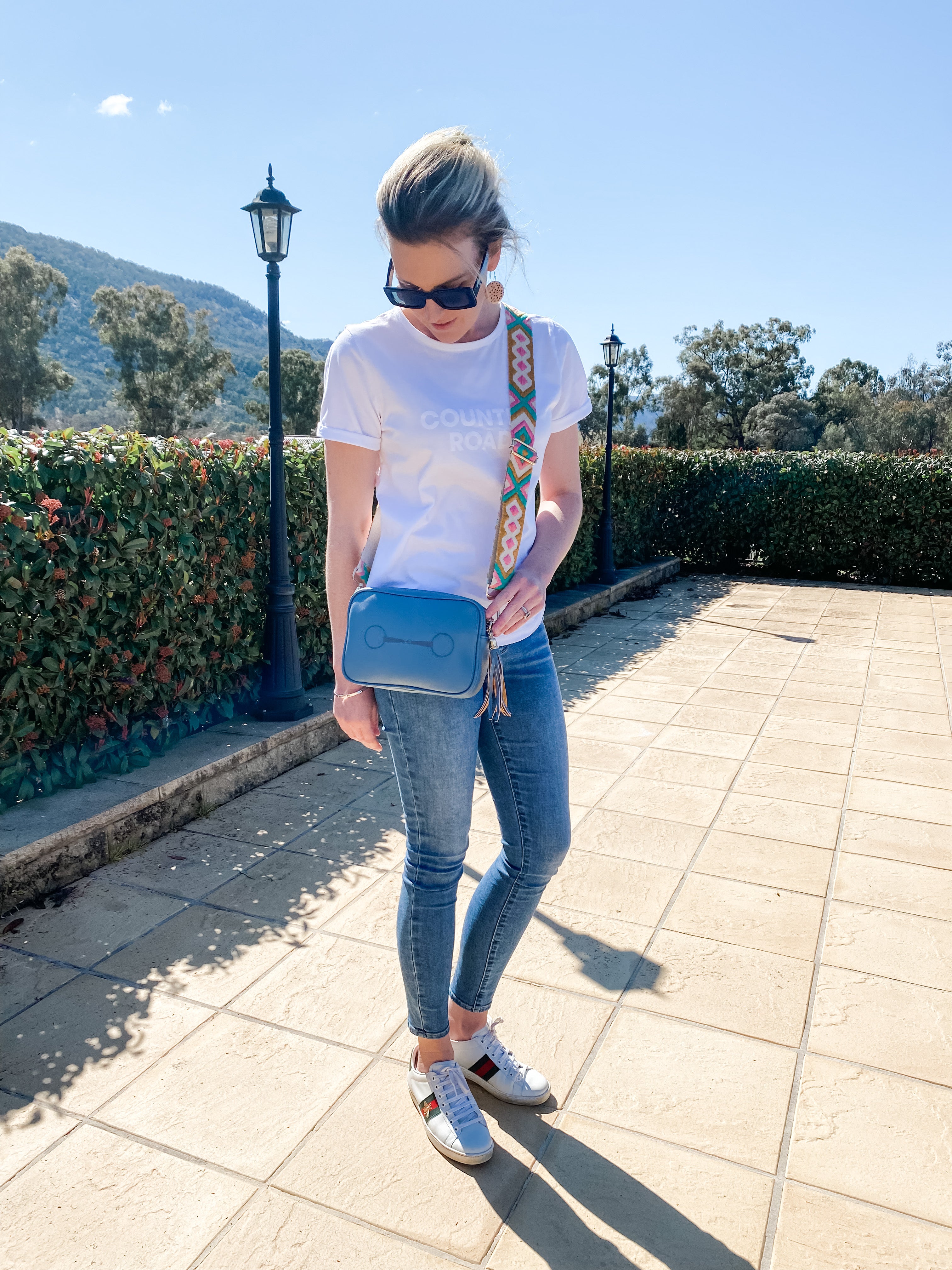 A blue handbag with a horsebit logo and aztec strap shown being worn by a lady in jeans and a white t shirt standing in front of a hedge